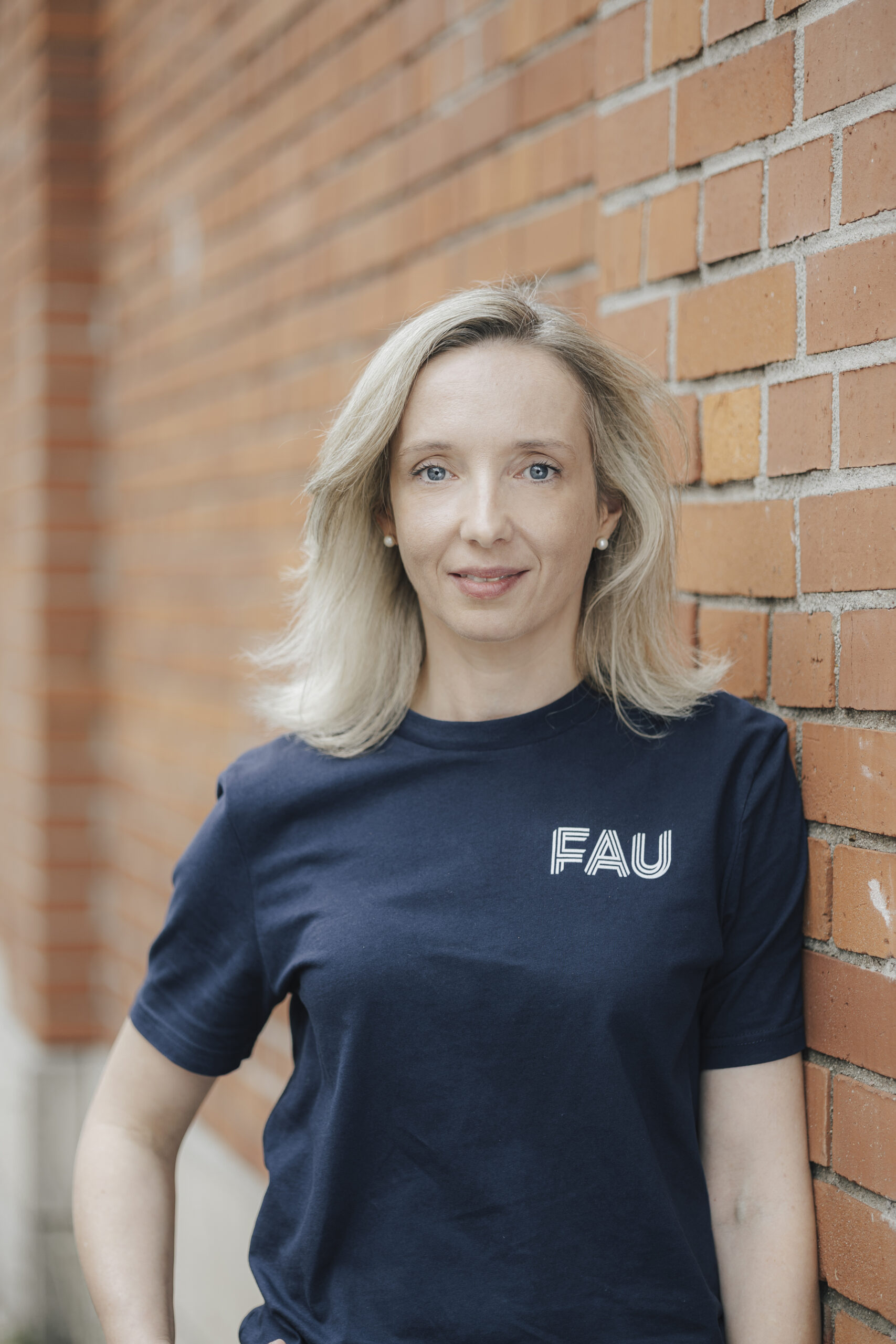 Portrait von Prof. Kathrin Castiglione (weiblich) an einer Ziegelwand im dunkelblauem FAU T-Shirt